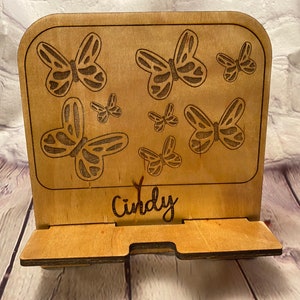 May include: A wooden phone stand with a butterfly design. The stand is made of light-colored wood and features an engraved pattern of butterflies of various sizes. The name "Cindy" is also engraved on the stand.