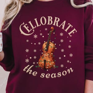 Peut inclure: Sweat-shirt bordeaux avec un motif festif. Le graphisme représente un violon orné de lumières, de flocons de neige et les mots "Cellobrate the season". Une tasse avec des bonhommes en pain d'épices est tenue à la main.