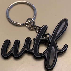 May include: A metal keychain with the letters "wtf" in a stylized, cursive font. The letters are black with a silver outline and are attached to a silver key ring.