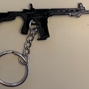 Puede incluir: Un llavero de metal negro con forma de rifle. El llavero tiene una cadena plateada y un llavero plateado. El rifle está detallado con varias partes y características. El diseño general es elegante y moderno.