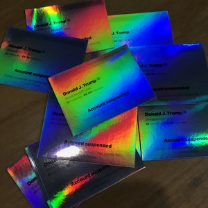 May include: A collection of holographic cards with the text "Donald J. Trump" and "Account suspended." The cards have a rainbow sheen and feature Twitter account details. The cards are scattered on a wooden surface.