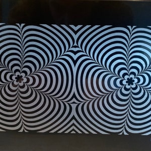 May include: A black and white patterned metal license plate with a swirling design. The design features two sets of concentric circles that create a hypnotic effect.