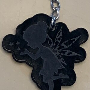 Könnte beinhalten: Schwarzer Schlüsselanhänger mit einer Silhouette einer Fee mit einem blattförmigen Flügel. Die Fee bläst auf einen Löwenzahn, und drei kleine Löwenzahnsamen schweben in der Luft.