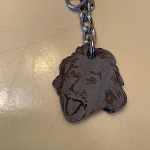 Pode incluir: Um chaveiro de prata com um amuleto de metal que apresenta uma imagem em preto e branco de Albert Einstein mostrando a língua.