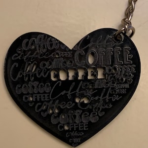 Op de afbeelding: Een zwarte hartvormige sleutelhanger met het woord "COFFEE" meerdere keren geschreven in verschillende lettertypen en formaten.