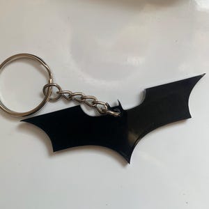 Pode incluir: Um chaveiro preto com o logotipo do Batman. O chaveiro apresenta uma corrente prateada e um grande símbolo de morcego estilizado. O símbolo do morcego é de cor preta sólida e tem um acabamento brilhante. O anel do chaveiro é prateado.