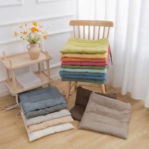 Puede incluir: Una colección de cojines cuadrados para asiento en varios colores sólidos, incluyendo verde lima, amarillo, naranja, rosa, turquesa, azul, azul oscuro, gris, beige y marrón. Los cojines presentan un acabado de tela texturizada y líneas cosidas.