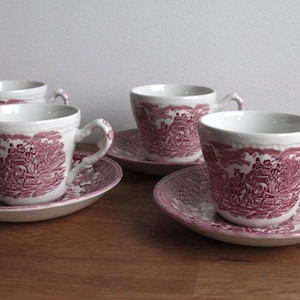 Puede incluir: Cuatro tazas y platillos blancos con un patrón de toile rojo. Las tazas tienen asas curvas y se asientan sobre platillos a juego. El diseño presenta una escena rural. El juego está dispuesto sobre una superficie de madera.