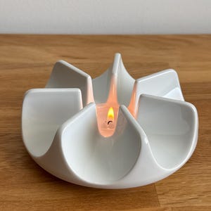 Könnte beinhalten: Ein weißer Kerzenhalter aus Keramik mit einem blütenartigen Design. Der Halter hat sechs gebogene Abschnitte, jeder mit einem erhöhten Rand. In der Mitte befindet sich eine brennende Kerze, die ein warmes Licht wirft. Der Halter steht auf einer Holzoberfläche.