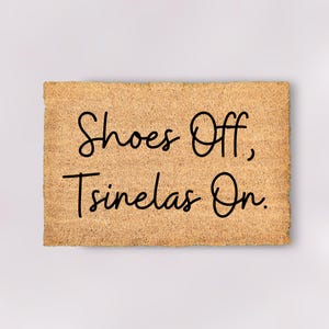 May include: A rectangular doormat made from natural coir fibre. The mat displays the text "Shoes Off, Tsinelas On" in black, cursive script. The coir has a light brown hue, and the text is centrally positioned.