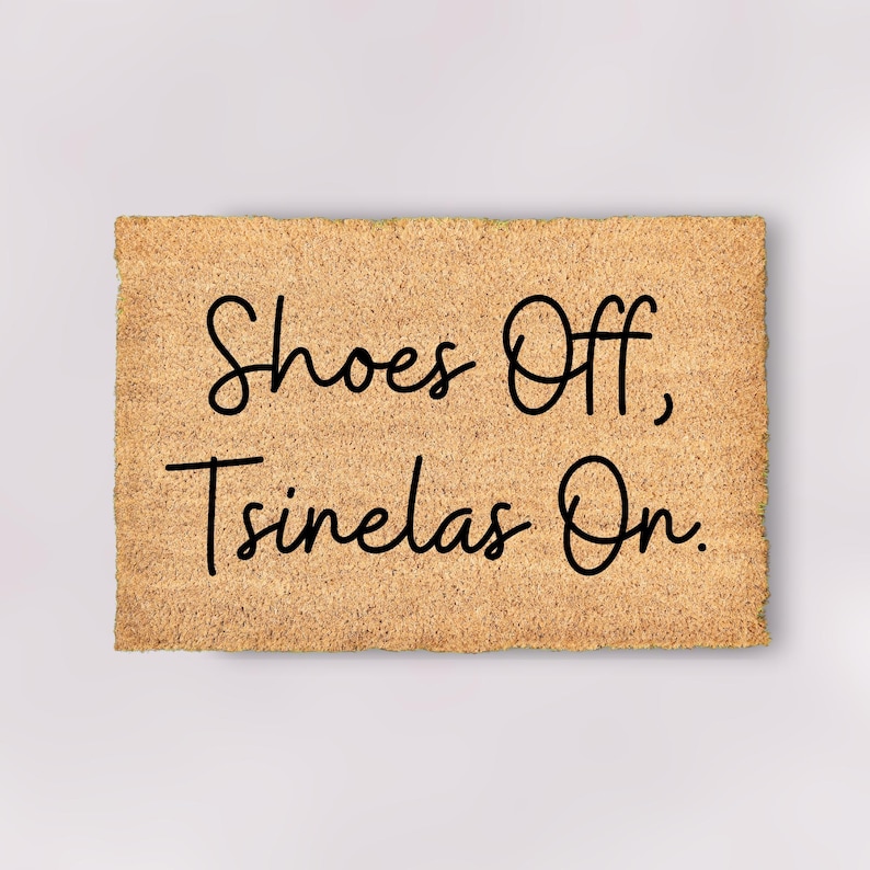 May include: A rectangular doormat made from natural coir fibre. The mat displays the text "Shoes Off, Tsinelas On" in black, cursive script. The coir has a light brown hue, and the text is centrally positioned.