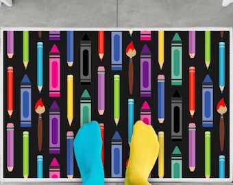 May include: A black and white doormat with a repeating pattern of colorful crayons and paintbrushes. The doormat has a white border.