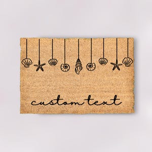 May include: A brown coir doormat with a design of seashells and starfish hanging from strings. The text "custom text" is printed on the doormat.