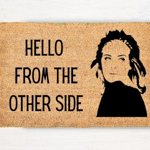 May include: A brown coir doormat with the text "HELLO FROM THE OTHER SIDE" in black lettering. A black silhouette of a woman's face is on the right side of the mat.