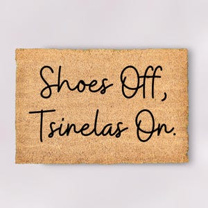 May include: A rectangular doormat with a light brown, textured surface. The mat features the phrase "Shoes Off, Tsinelas On" in a black, cursive font. The doormat is designed for an entryway.