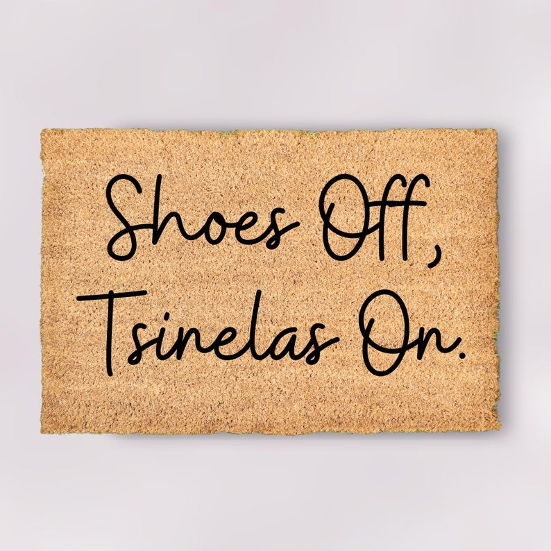 May include: A rectangular doormat with a light brown, textured surface. The mat features the phrase "Shoes Off, Tsinelas On" in a black, cursive font. The doormat is designed for an entryway.