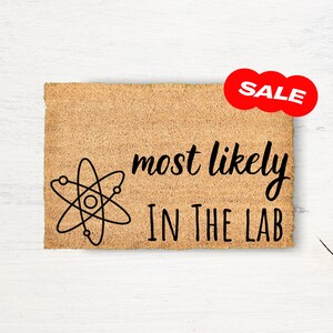 May include: A brown doormat with the text "most likely in the lab" and a stylized atom symbol. The word "SALE" is in a red circle above the text.
