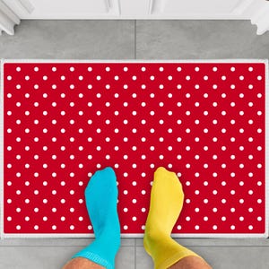 May include: A red doormat with white polka dots. Two feet are standing on the doormat, one wearing blue socks and the other wearing yellow socks.