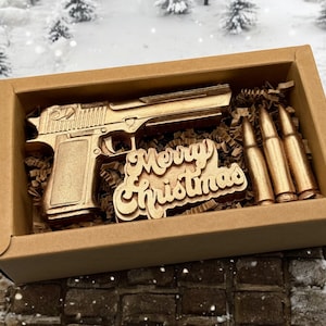 Pode incluir: Uma caixa de presente dourada com uma pistola dourada, balas e um letreiro de "Feliz Natal" dentro. A caixa está sobre uma superfície de tijolo coberta de neve.