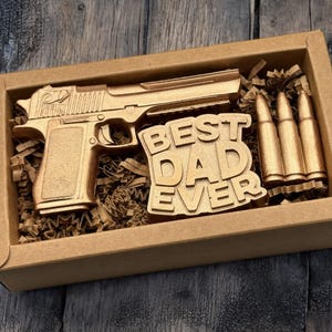 May include: A gold-colored gift set in a cardboard box. The set includes a replica handgun, three bullets, and a sign that reads "BEST DAD EVER". The items are nestled in brown paper filler, creating a unique gift.