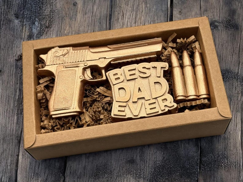 May include: A gold-colored gift set in a cardboard box. The set includes a replica handgun, three bullets, and a sign that reads "BEST DAD EVER". The items are nestled in brown paper filler, creating a unique gift.