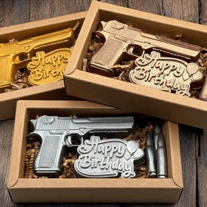 Puede incluir: Tres cajas de regalo con objetos en forma de pistola en oro y plata. Cada caja contiene una pistola, un letrero de "Feliz Cumpleaños" y dos balas. Sobre una superficie de madera.