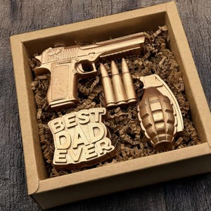 May include: A wooden gift box containing a gold-colored toy pistol, three bullets, a grenade, and a sign that reads "BEST DAD EVER". The items are nestled in brown paper filler.