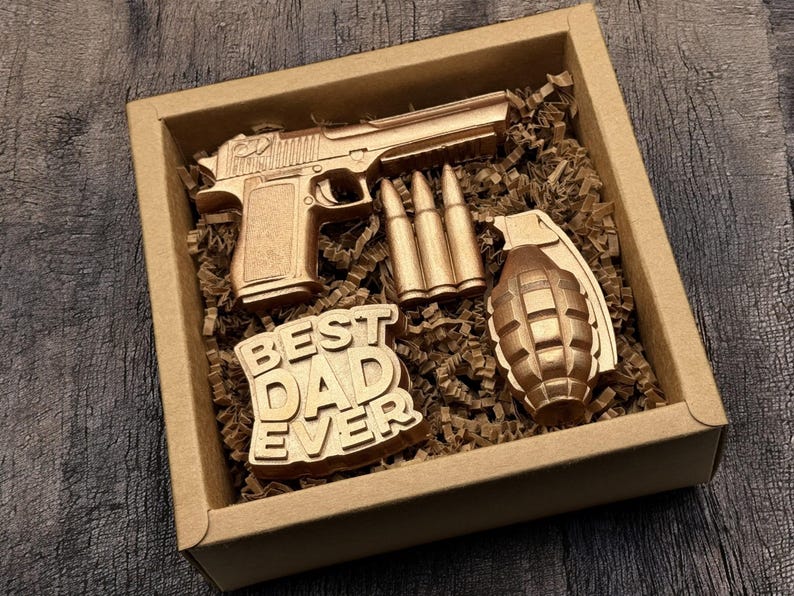 May include: A wooden gift box containing a gold-colored toy pistol, three bullets, a grenade, and a sign that reads "BEST DAD EVER". The items are nestled in brown paper filler.
