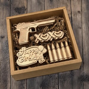 May include: A gold-colored gift box containing a gun, the letters "XOXO", bullets, and a "Happy Birthday" message. The items are nestled in brown paper and set against a wooden background.