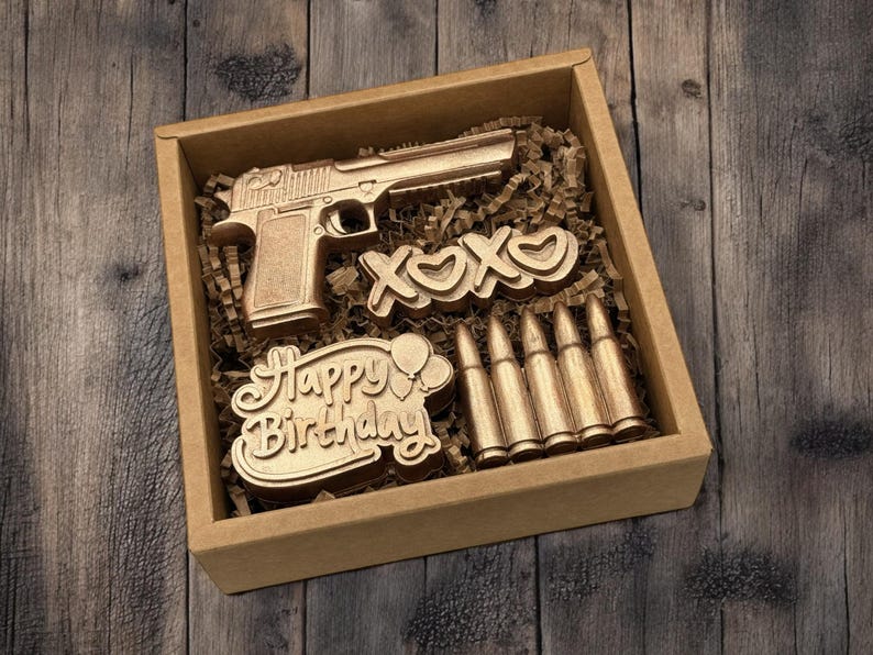 May include: A gold-colored gift box containing a gun, the letters "XOXO", bullets, and a "Happy Birthday" message. The items are nestled in brown paper and set against a wooden background.