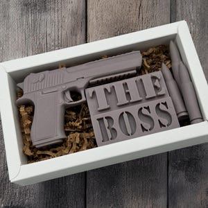Pode incluir: Uma caixa de presente branca com uma arma marrom, duas balas marrons e uma barra de sabão marrom que diz "THE BOSS" dentro.