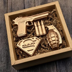 May include: A gift box containing a gold-colored pistol, three bullets, a grenade, and a Superman logo with the word "DAD" inside. The items are nestled in brown paper shreds within a cardboard box.