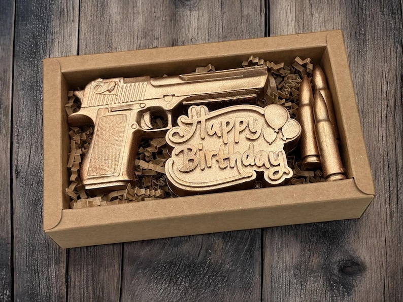 May include: A gold-colored decorative gift set in a cardboard box. The set includes a pistol, two bullets, and a "Happy Birthday" sign with balloons. The items are nestled in brown paper shreds, creating a unique birthday gift.
