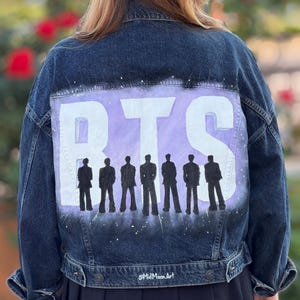 Può includere: Giacca di jeans blu scuro con un disegno dipinto sul retro. Il disegno presenta le lettere "BTS" in bianco su uno sfondo viola, con sagome di sette figure sottostanti. La firma dell'artista è in basso.