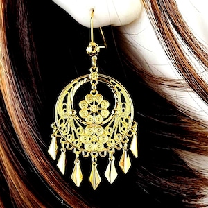 May include: Gold-toned filigree earrings with a circular design and dangling diamond-shaped charms. The earrings feature intricate floral and geometric patterns, and are shown against a backdrop of brown hair.