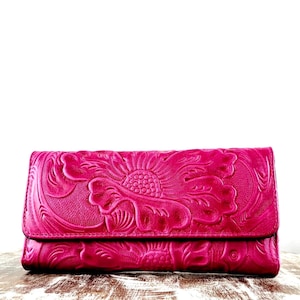 May include: A rectangular hot pink wallet featuring an embossed floral pattern across the entire front surface. The wallet has a flap closure that extends across the width of the item, showcasing a detailed, raised texture of large flower petals.