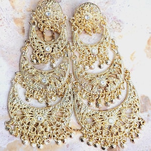 May include: A pair of ornate gold-colored earrings. Each earring features a circular top with a central gem, connected to three crescent-shaped layers with intricate filigree and dangling beads. The earrings are designed with a vintage aesthetic.