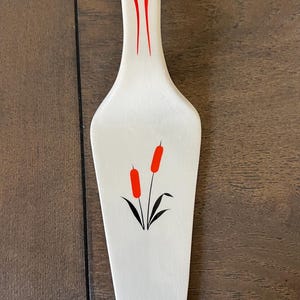May include: A vintage white ceramic pie server with a decorative red and black cattail design. The handle features red accent lines. This kitchen utensil is ideal for serving pies, cakes, and other baked goods.