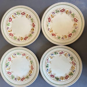 Puede incluir: Cuatro platos de cerámica blanca con un diseño floral. Cada plato presenta una corona floral central en rojo, morado y amarillo, rodeada por un borde verde y beige. Los platos están dispuestos sobre una superficie gris.