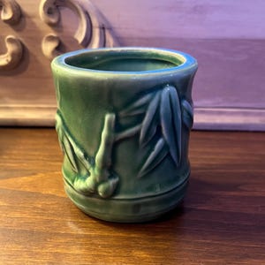 Pode incluir: Uma pequena chávena cilíndrica de cerâmica verde com um desenho de bambu. A chávena tem um acabamento brilhante e uma superfície ligeiramente texturizada. O desenho de bambu é em relevo. A chávena tem cerca de 7,6 cm de altura.