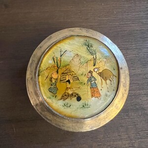 May include: Circular antique metal trinket box with a painted scene. The lid features a detailed miniature painting of figures, a tent, and a camel in a desert landscape. The box has a gold-coloured metal rim and a light yellow interior.
