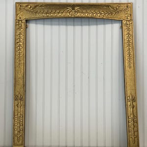 Puede incluir: Un marco de chimenea rectangular de metal color dorado con un patrón en relieve ornamentado. El marco incluye motivos florales decorativos, cintas onduladas y columnas verticales con detalles de remate en la base.