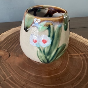 May include: A ceramic, hand-painted vase with a beige base and a brown and green rim. The vase features a floral design with white flowers and red centers, surrounded by green leaves. The vase is displayed on a wooden surface.