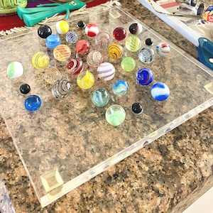 May include: A square, clear acrylic game board featuring a grid of indentations, each holding a spherical glass marble. The marbles vary in size, color, and pattern, including solid, swirled, and striped designs in blue, red, yellow, green, and black.