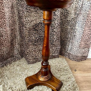 May include: A dark brown, antique wooden plant stand with a round top and a pedestal base. The stand has a three-legged base and a tall, slender stem. The wood has a glossy finish, and the stand is likely used for displaying plants or decorative items.