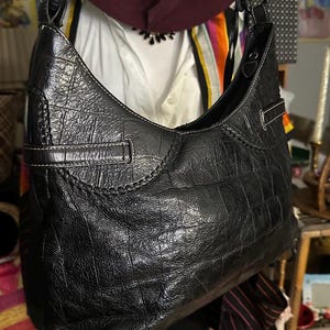Puede incluir: Bolso de hombro de cuero negro con textura de cocodrilo. El bolso tiene forma curva, una sola correa para el hombro y detalles de costura blancos. Una hebilla plateada es visible en el lateral. Un accesorio elegante.