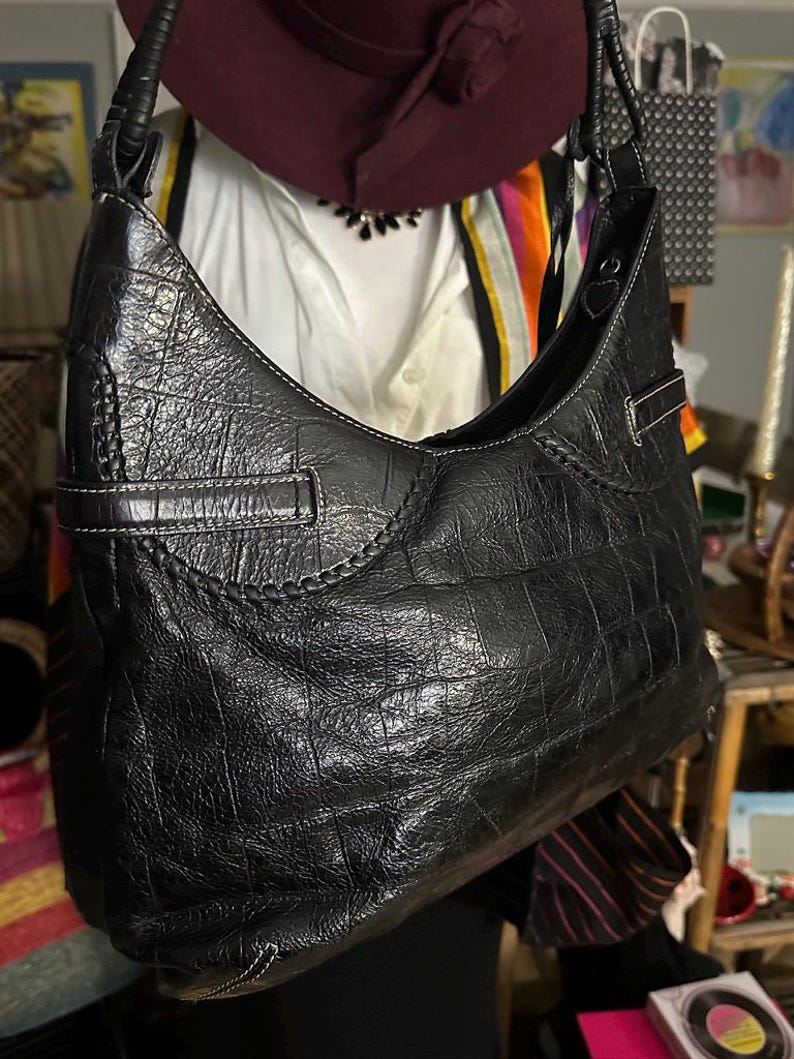 Puede incluir: Bolso de hombro de cuero negro con textura de cocodrilo. El bolso tiene forma curva, una sola correa para el hombro y detalles de costura blancos. Una hebilla plateada es visible en el lateral. Un accesorio elegante.