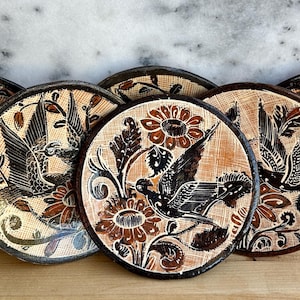 May include: A collection of six round ceramic plates with a hand-painted design. Each plate features a central bird motif in black and brown, surrounded by floral and leaf patterns. The plates have a textured, earthy appearance.