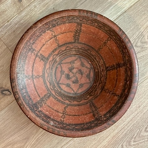 May include: A large, round, terracotta bowl with a black geometric design. The bowl has a wide rim and a deep, rounded interior. The design features a central circle with a series of concentric circles and lines radiating outward.