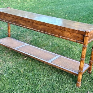 May include: A long, rectangular console table with a smooth, medium-brown wood top and a lower shelf featuring three woven cane panels. The frame is constructed with a faux bamboo design, featuring segmented vertical legs and horizontal supports.